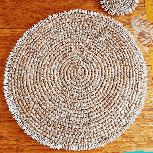 Individual redondo de rafia natural con borde de conchitas estilo boho sobre mesa.